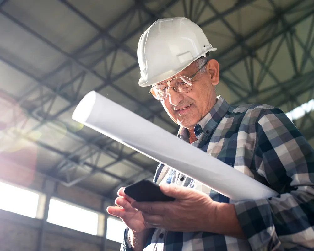 Contractor in a hard hat holding rolled plans and a phone on a job site